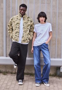 Fleecejacka med kamouflagemönster i beige och grönt, tillsammans med en vit T-shirt, svarta vida jeans och svartvita sneakers.