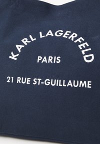 Saco de tecido azul-marinho com texto branco: "KARL LAGERFELD PARAIS 21 RUE ST-GUILLAUME" numa fonte arredondada. Design robusto.
