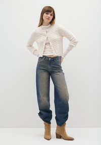 Off-white kabelgebreid vest met knopen, gedragen over een kanten top. Donkerblauwe wijd uitlopende jeans met een vervaagde afwerking en beige enkellaarzen.