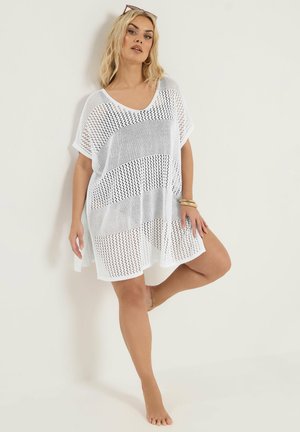 Kvinde med blondt hår iført hvid, strikket, løstsiddende kjole, stående barfodet mod en hvid væg, med det ene ben bøjet, iført solbriller på hovedet og guldbøjler.