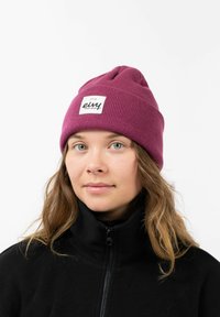 Purple knitted beanie with a folded cuff, featuring a square white label with black text. Worn over a black zip-up fleece jacket.