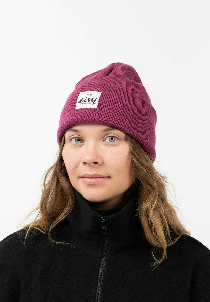 Purple knitted beanie with a folded cuff, featuring a square white label with black text. Worn over a black zip-up fleece jacket.