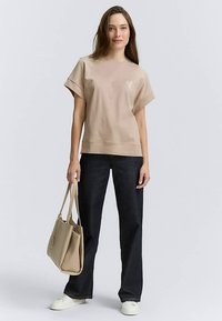 T-shirt beige oversize à manches courtes avec détails cousus, associé à un pantalon en denim foncé et un sac cabas beige clair. Des baskets blanches complètent le look.