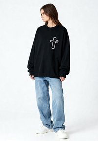 Femme portant un sweat-shirt noir oversize avec une croix blanche et le texte « KARMA KILLS », un jean bleu clair ample et des baskets blanches, debout de profil.