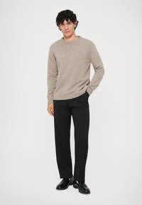 Μπεζ πλεκτό πουλόβερ με στρογγυλή λαιμόκοψη, συνδυασμένο με κομψά μαύρα παντελόνια και μαύρα παπούτσια, φορεμένο από ένα μοντέλο με σγουρά μαλλιά.