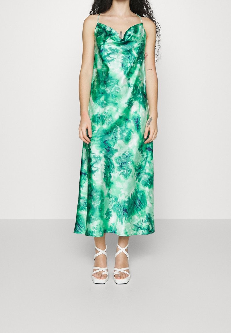 Femme portant une robe nuisette en satin tie-dye verte et blanche à fines bretelles, accompagnée de sandales blanches à talons hauts à lanières, se tenant devant un fond uni.