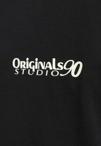 Crna pamučna majica s bijelim natpisom "Originals90 STUDIO." Natpis je podebljan, centriran i malo zakrivljen. Glatka tekstura.