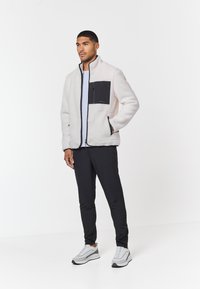 Fleecová bunda v off-white farbe s čiernou prednou vreckom, so zipsom a bočnými vreckami; skombinovaná s čiernymi nohavicami a šedými teniskami.