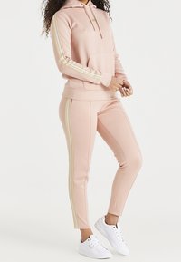 Roze hoodie en bijpassende broek van zacht materiaal, met gouden strepen langs de zijkanten en een zak aan de voorkant. Witte sneakers maken de outfit compleet.