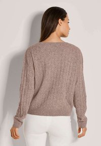 Vrouw met lang donker haar, gekleed in een mauve kabelgebreide trui en witte broek, met haar rug naar de camera gekeerd tegen een effen lichte achtergrond.