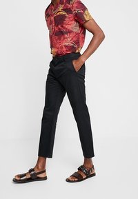 Camicia a maniche corte con un motivo di foglie rosse e gialle, pantaloni slim-fit neri e sandali in pelle marrone con dettagli a fibbia.