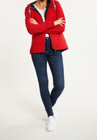 Personne portant un sweat à capuche rouge zippé, une chemise blanche, un jean skinny bleu foncé et des baskets blanches, se tenant les bras croisés avec une jambe fléchie.