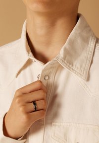 Witte denimjacket met een geknopt kraag en zichtbare stiksels. Hand houdt een zwarte ring bij de halslijn, textuur van de stof is duidelijk zichtbaar.