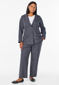 Traje gris de rayas con un blazer ajustado que tiene dos bolsillos frontales y pantalones a juego. Combinado con una blusa blanca y unos zapatos planos negros.