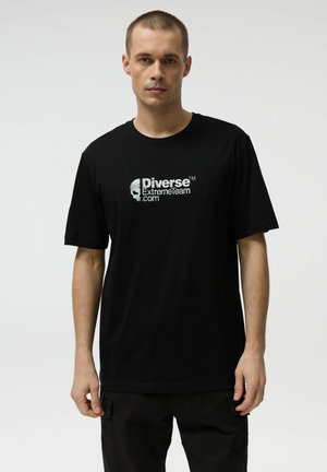 Mężczyzna o krótkich włosach, ubrany w czarną koszulkę z białym logo "Diverse ExtremeTeam.com", stojący na jednolitym jasnym tle.