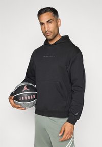 Μαύρη φούτερ μπλούζα με κουκούλα και μπροστά τσέπη, με διακριτό λογότυπο "Jordan Sports". Το μοντέλο κρατάει μια μπάλα μπάσκετ με υφή.