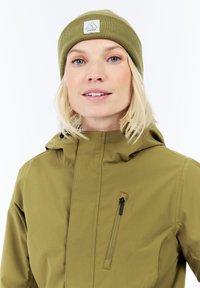 Žena so svetlými blond vlasmi, ktorá má na sebe olivovo zelenú čiapku a zodpovedajúcu bundu s vreckom na zips na hrudi, sa jemne usmieva na bielom pozadí.