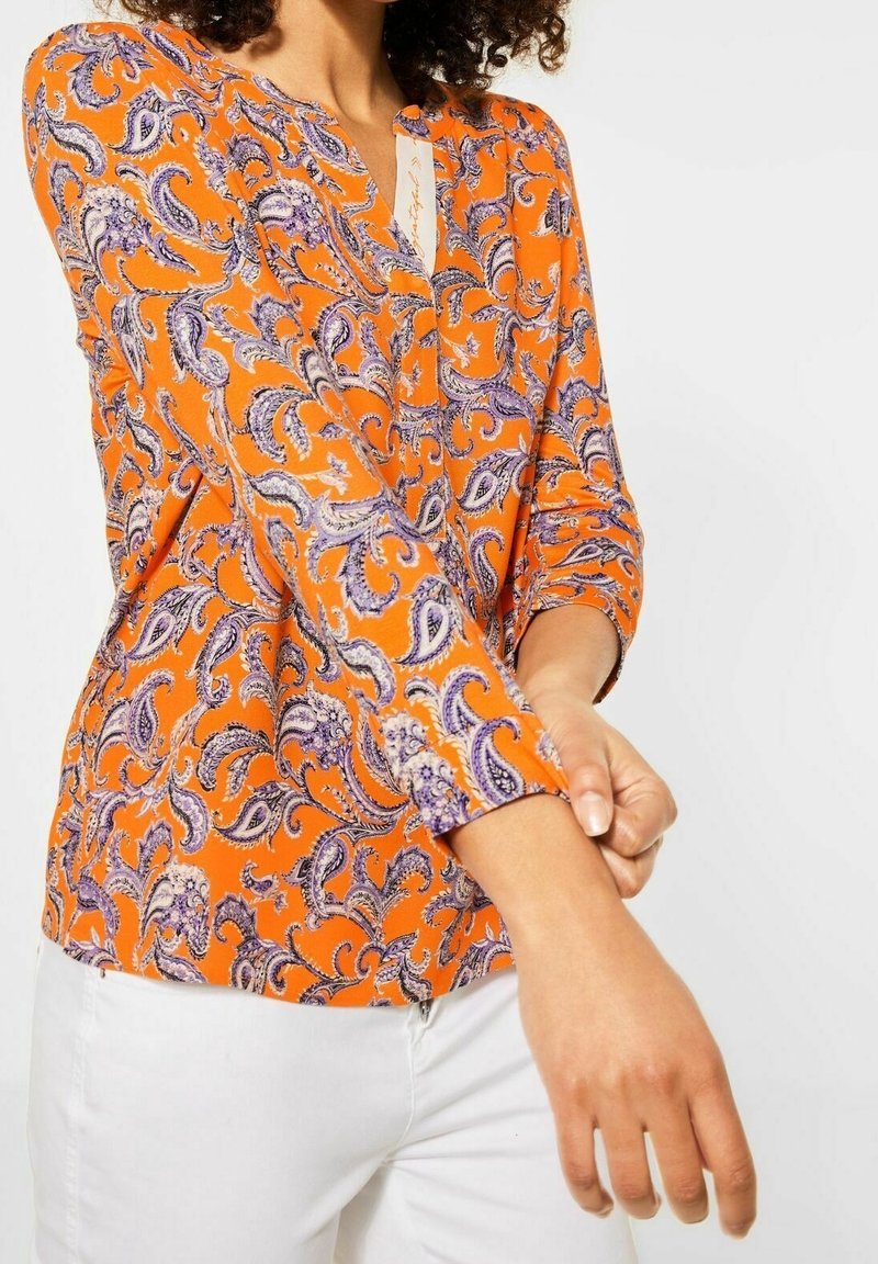 Blouse à motif cachemire en orange avec des accents violets. Manches longues avec poignets et encolure en V. Tissu léger à texture lisse.
