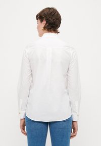 Hvitt skjorte laget av bomull med lange ermer, med bakfelling og gullknappeaksenter ved mansjettene, vist med blå jeans.