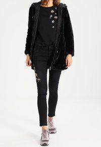 Svart fuskpälsjacka med huva, dragkedjeslutning och broderade blommönster. Bärs med en svart skjorta, svarta skinnyjeans och ljuslila sneakers.