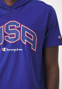 Niebieska koszulka z krótkim rękawem i kapturem, z wyraźnym napisem "USA" w czerwonych i białych obrysowanych literach; logo "Champion" w białym kolorze nad dołem.