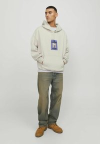 Lys grå hættetrøje med en frontlomme, prydet med et blåt grafisk design. Sammen med bleget blå jeans og brune snørebåndssko.