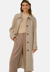 Beige wollen blend jas met een kraag, lange mouwen en vijf bruine knopen. Gecombineerd met een lichtbeige coltrui en wijde beige broek.
