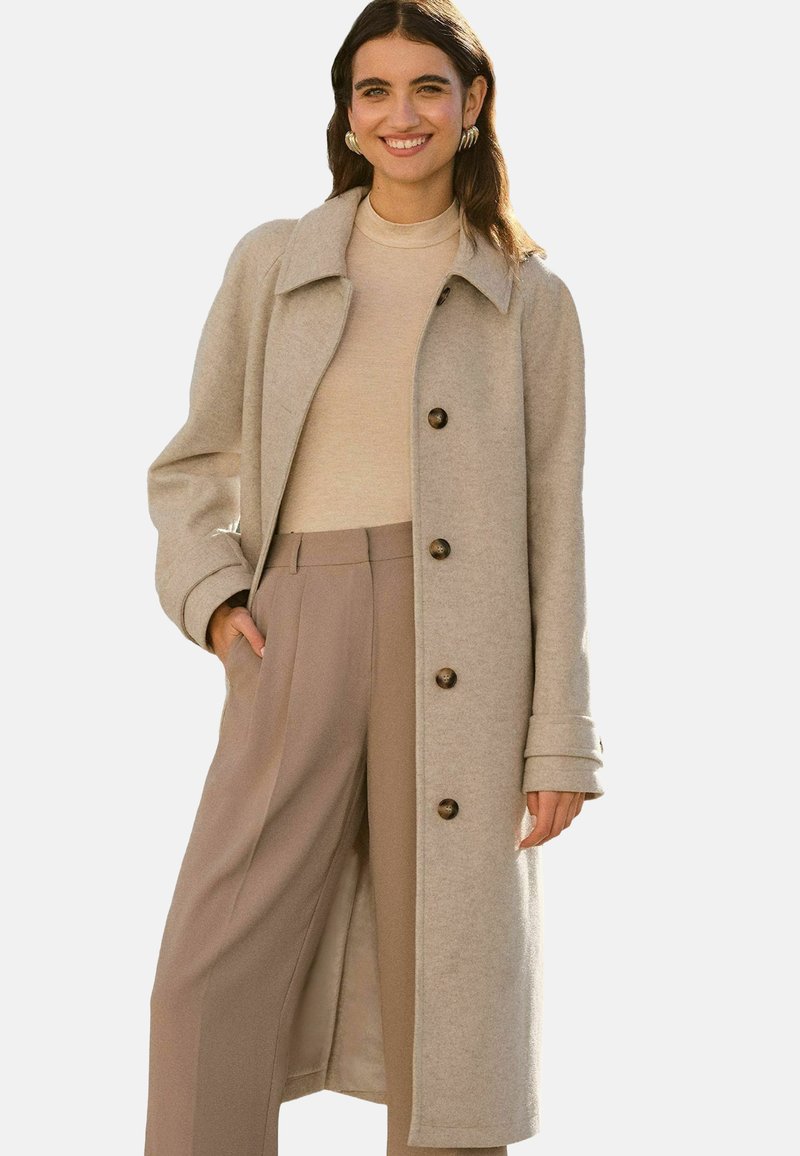 Beige wollen blend jas met een kraag, lange mouwen en vijf bruine knopen. Gecombineerd met een lichtbeige coltrui en wijde beige broek.