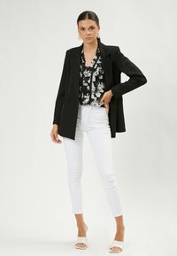 Schwarze Blazer über einer blumigen Bluse, kombiniert mit weißen Skinny-Jeans und weißen Rutsch-Sandalen. Das Modell steht vor einem neutralen Hintergrund.