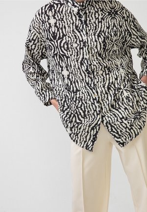 Chemise boutonnée à motifs abstraits en noir et blanc portée avec un pantalon beige clair, mains dans les poches, sur un fond uni.