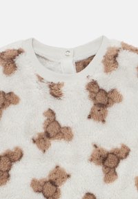 Material alb moale și pufos cu un model de ursuleț maro și un decolteu rotund și nervurat cu închidere cu nasture tip capse în spate.