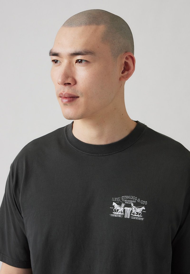 Camiseta de algodón gris oscuro con un estampado gráfico blanco en el lado izquierdo del pecho, mangas cortas y cuello redondo. Textura suave.