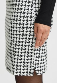 Houndstooth blyantskørt i sort og hvidt med en sideslids. Har et struktureret stof og tætsiddende pasform. Hånden hviler mod stoffet.
