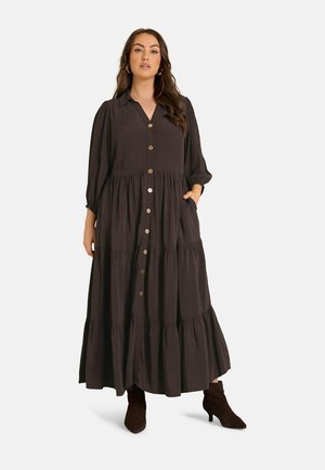 Donna che indossa un lungo vestito marrone scuro con bottoni, gonna a balze e maniche a tre quarti, abbinato a stivaletti scuri, in piedi con la mano in tasca.