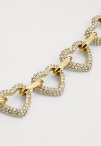Auksinis apyrankė, kurioje yra širdies formos grandinės, puošti skaidriais rhinestone akmenimis ir sujungti mažomis aukso užsegimais, pažymėta "GUESS."