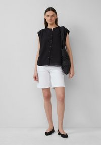 Blouse noire à manches courtes avec boutons, shorts blancs jusqu'aux genoux et chaussures à talons ouverts noires. Un sac fourre-tout noir texturé est porté sur l'épaule.