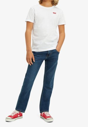 Enfant portant un t-shirt blanc Levi's, un jean bleu et des baskets rouges avec des lacets blancs, debout avec une main dans la poche et l'autre le long du corps.