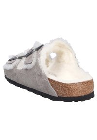 Pantofole grigie a infilare con tomaia in suede, fodera bianca pelosa, intersuola in sughero e suola in gomma nera; dotate di due fibbie metalliche regolabili.