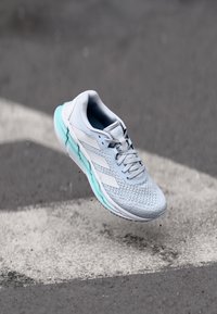 Shoe sportiv de un albastru deschis, cu un superior texturat, accente albe și o talpă căptușită, de culoare albastră, surprins în aer, pe un fundal gri.