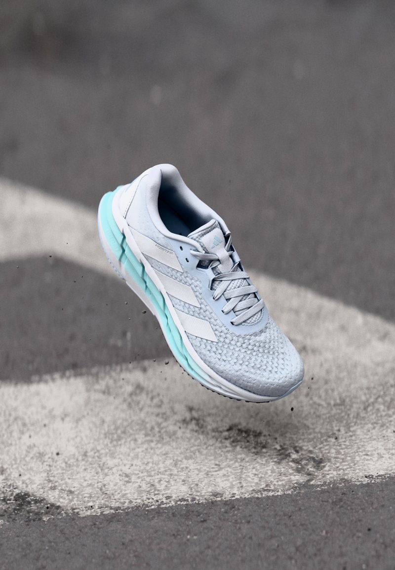 Zapatilla deportiva de color azul claro con un upper texturizado, detalles en blanco y una suela azul acolchada, capturada en el aire sobre un fondo gris.