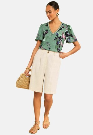 Kvinde iført en grøn blomsterprint V-hals bluse, beige højtaljede knælange shorts, guldbelagte øreringe, armbånd, med en vævet håndtaske og remsandal.