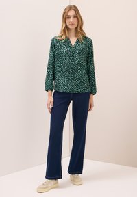 Vrouw die staat en een groen en zwart gebloemd blouse, donkerblauwe wijde jeans en beige sneakers draagt tegen een effen lichte achtergrond.