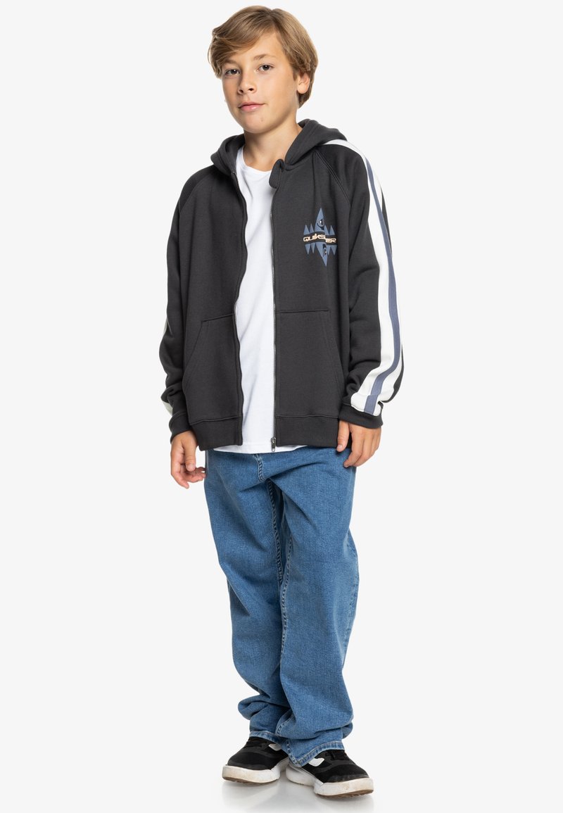 Sudadera negra con cremallera y dos bolsillos frontales, diseño de logo azul, rayas blancas en las mangas, llevada sobre una camiseta blanca y unos jeans de mezclilla.