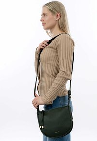 Kobieta z blond włosami, ubrana w beżowy sweter z prążkowanej dzianiny i niebieskie jeansy, trzyma czarną skórzaną torebkę na ramię z złotymi akcentami.