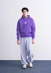Lilla hettegenser med lomme foran, kombinert med lyseblå oversized bukser og hvite sneakers, satt mot en ensfarget bakgrunn.