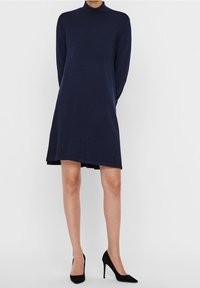 Marinblå klänning med långa ärmar och hög krage, med en lös passform och lätt utsvängd fåll, i kombination med svarta högklackade skor.
