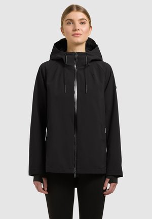 Junge Person mit zurückgebundenem Haar, die eine schwarze Kapuzenjacke mit Frontreißverschluss trägt und vor einem schlichten hellen Hintergrund steht.