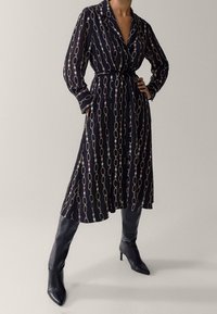 Pitkähihainen tummansininen mekko, jossa on valkoisia ja vaaleanpunaisia ketjumaisia kuvioita, vyötärövyö ja kaulus. Yhdistetty polven ylle ulottuviin mustiin korkokenkiin.