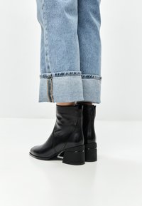Bottines en cuir noir avec un talon bloc, présentant une texture lisse et un design minimaliste, associées à un pantalon en denim bleu clair retroussé.
