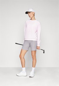 Ροζ υφασμάτινη μπλούζα, καλοκαιρινά σορτς με σχέδιο, λευκά αθλητικά παπούτσια και λευκό-μαύρο καπέλο. Κρατάει ένα μπαστούνι του γκολφ και στέκεται σε ουδέτερο φόντο.